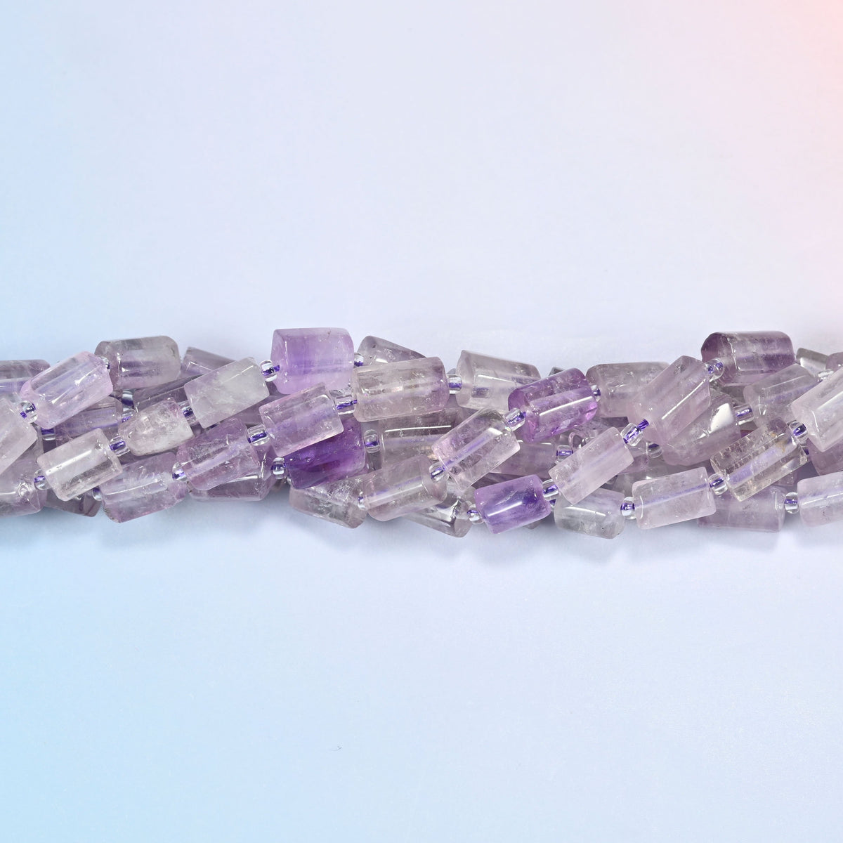 Lavender amethyst faceted tube beads crossing the frame in a shallow wave, several strands overlapping, cut faces catching light.