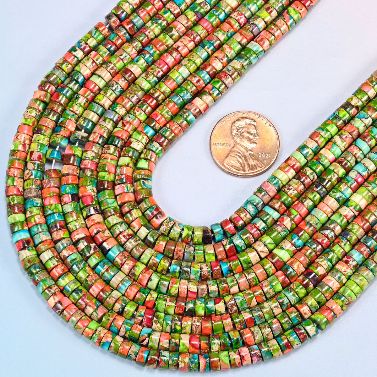 Close view of dyed blue green sea sediment jasper heishi disc strands curving beside a US penny for scale