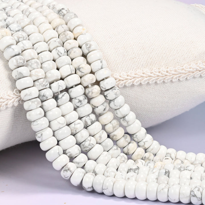 Close-up of multiple strands of polished White Howlite rondelle beads with gray marbled veining, draped over a cream fabric jewelry display bust.