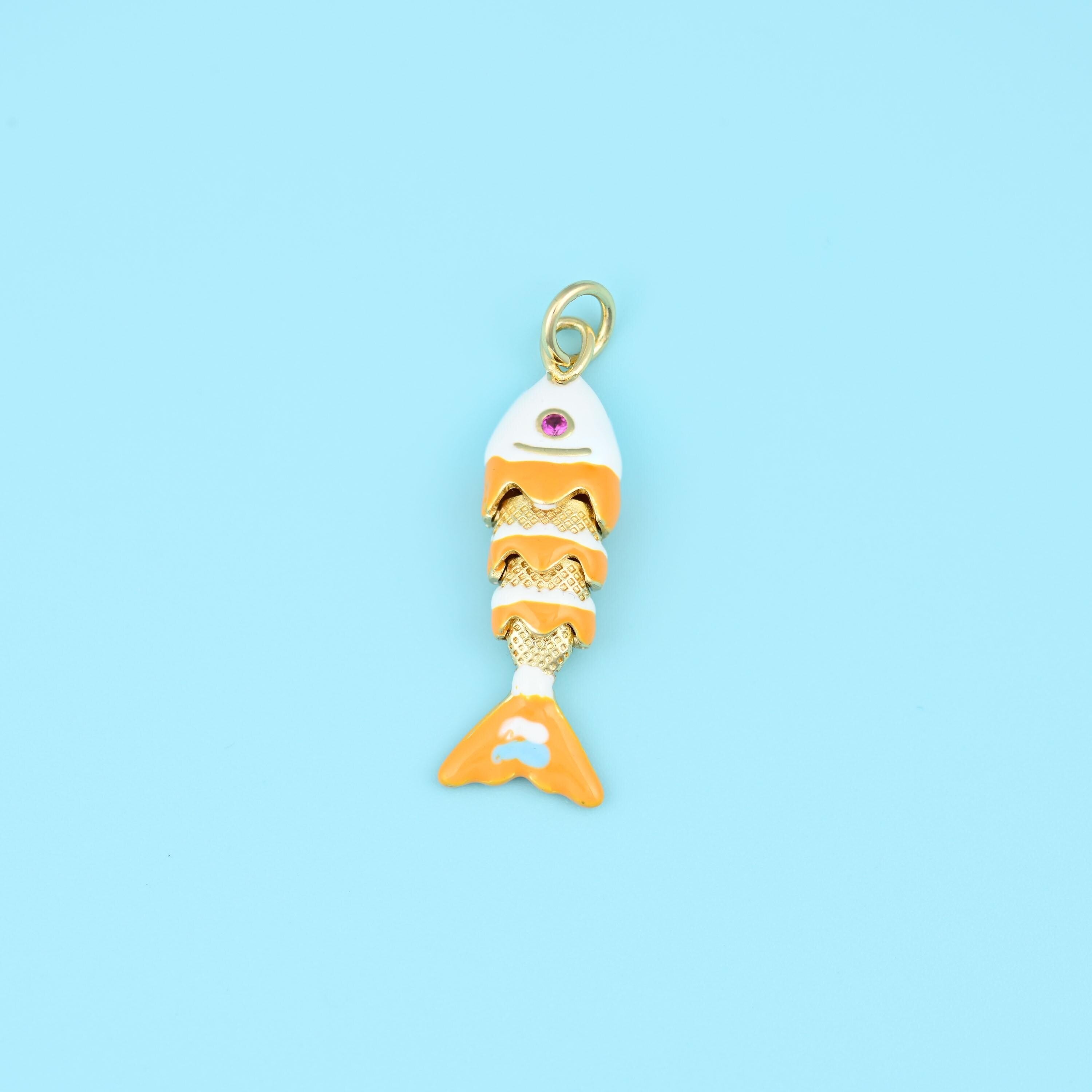 A jointed fish whose body segments swing as it hangs, finished in warm orange and white enamel over gold with a tiny pink…