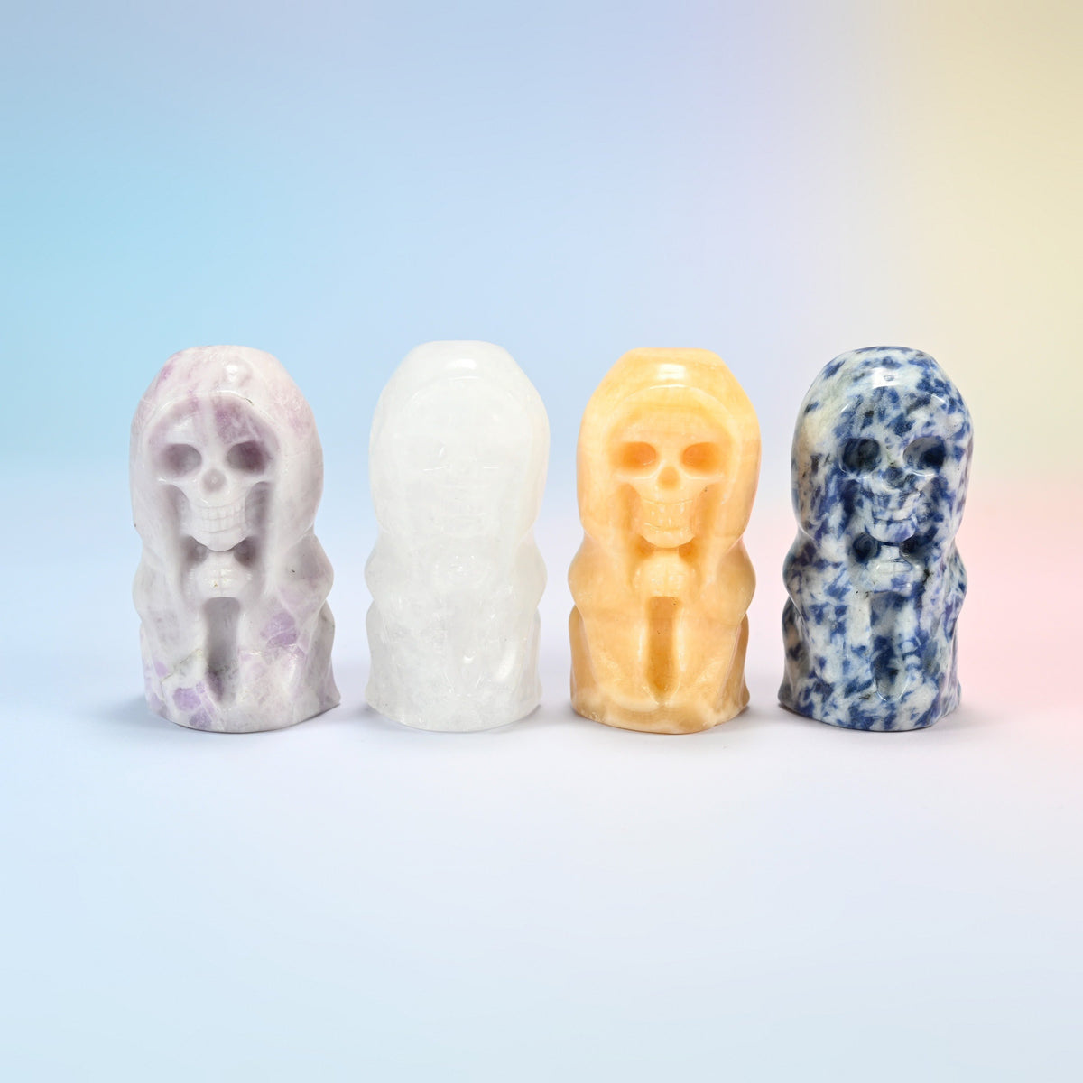 Four hooded skull carvings: natural lepidolite, clear quartz, yellow jade and blue spot jasper, hands clasped under the hood