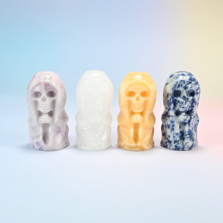 Four hooded skull carvings: natural lepidolite, clear quartz, yellow jade and blue spot jasper, hands clasped under the hood