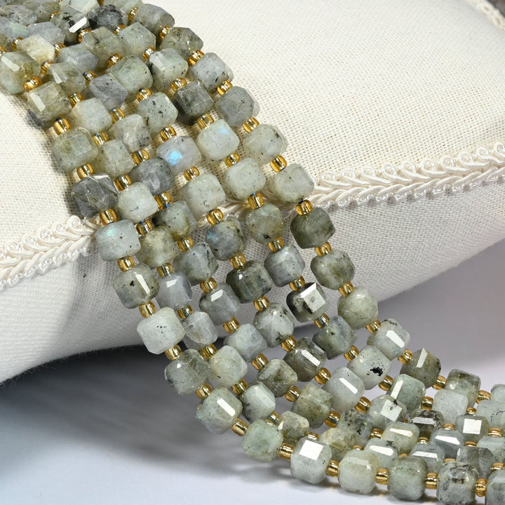 Gray labradorite faceted cube beads 6mm with small gold spacer beads, several strands draped over a white cushion