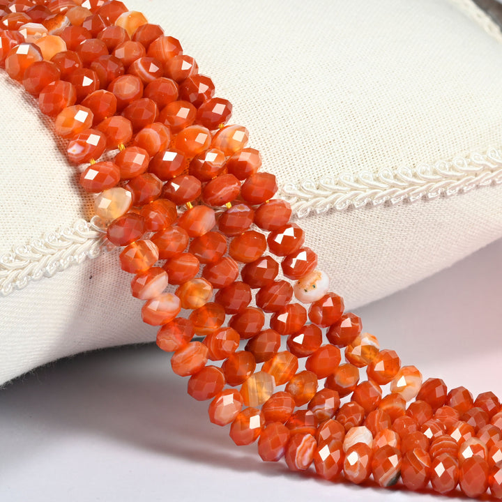 Dyed red strip agate faceted rondelle beads in warm orange red banded with white, three strands draped over a cream linen cushion with braided trim