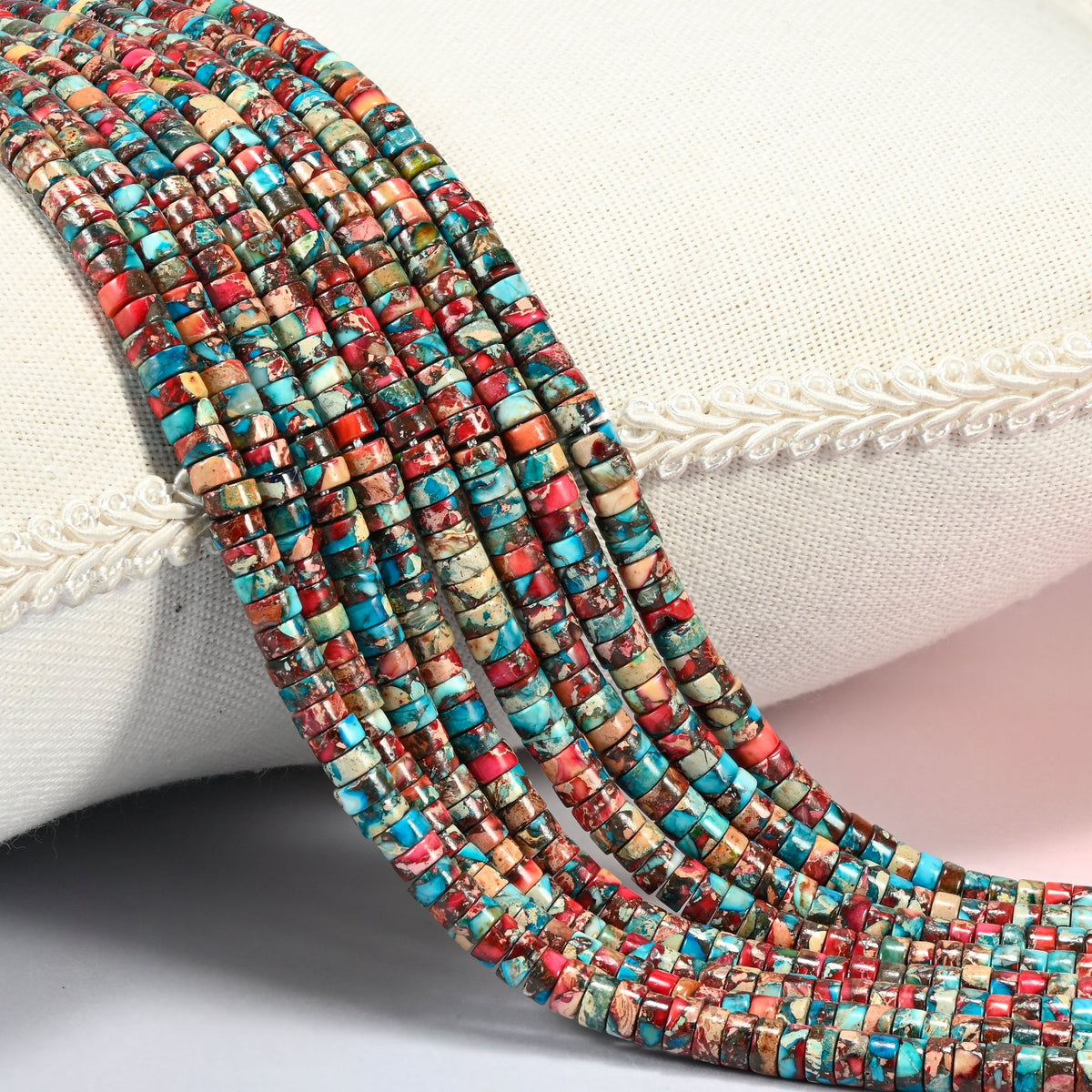 Dyed red blue sea sediment jasper heishi disc beads 2x4mm, several strands of turquoise, brick red and cream discs draped over a white cushion