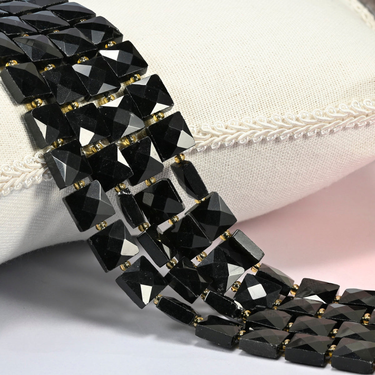 Natural black onyx faceted flat square beads 10x10mm with small gold seed bead spacers, several strands draped over a white cushion