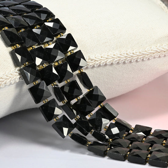 Natural black onyx faceted flat square beads 10x10mm with small gold seed bead spacers, several strands draped over a white cushion
