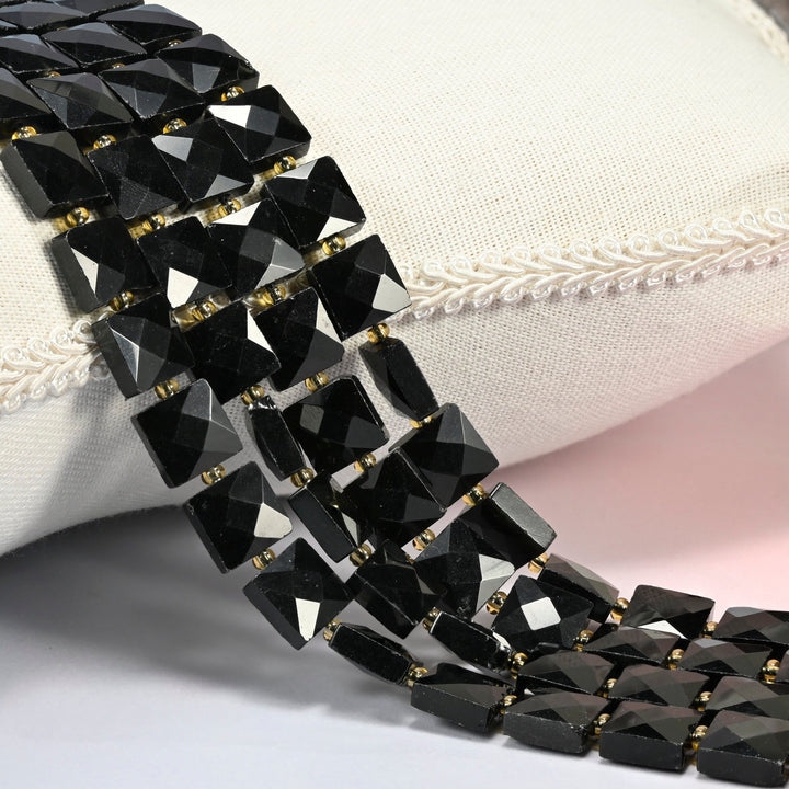 Natural black onyx faceted flat square beads 10x10mm with small gold seed bead spacers, several strands draped over a white cushion