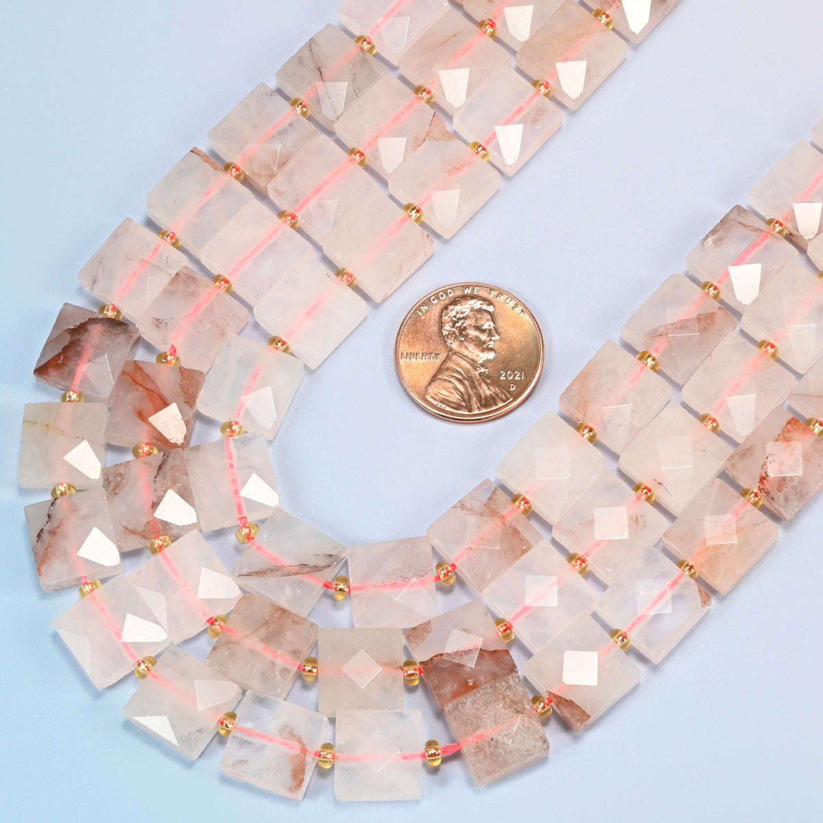 Close view of natural red hematoid quartz faceted flat square strands curving beside a US penny for scale