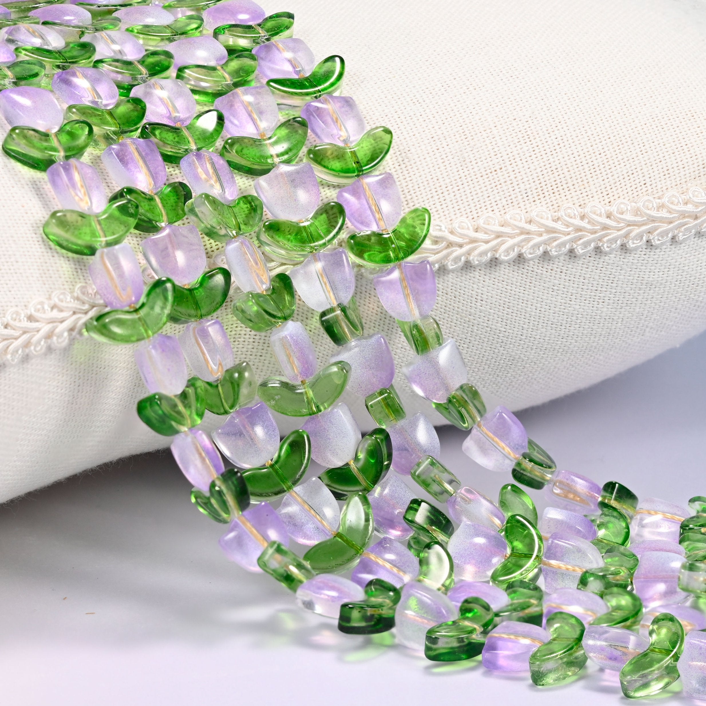 Close-up of lavender tulip flower and green leaf glass beads cascading diagonally over a cream woven pillow with decorative trim.