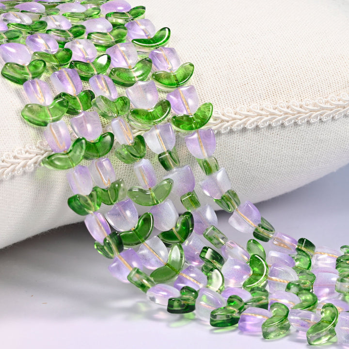 Close-up of lavender tulip flower and green leaf glass beads cascading diagonally over a cream woven pillow with decorative trim.