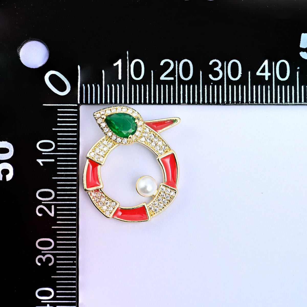 Round snake charm upright on a millimetre ruler, the red enamel ring and pearl set at the zero mark to show the width across the medallion.