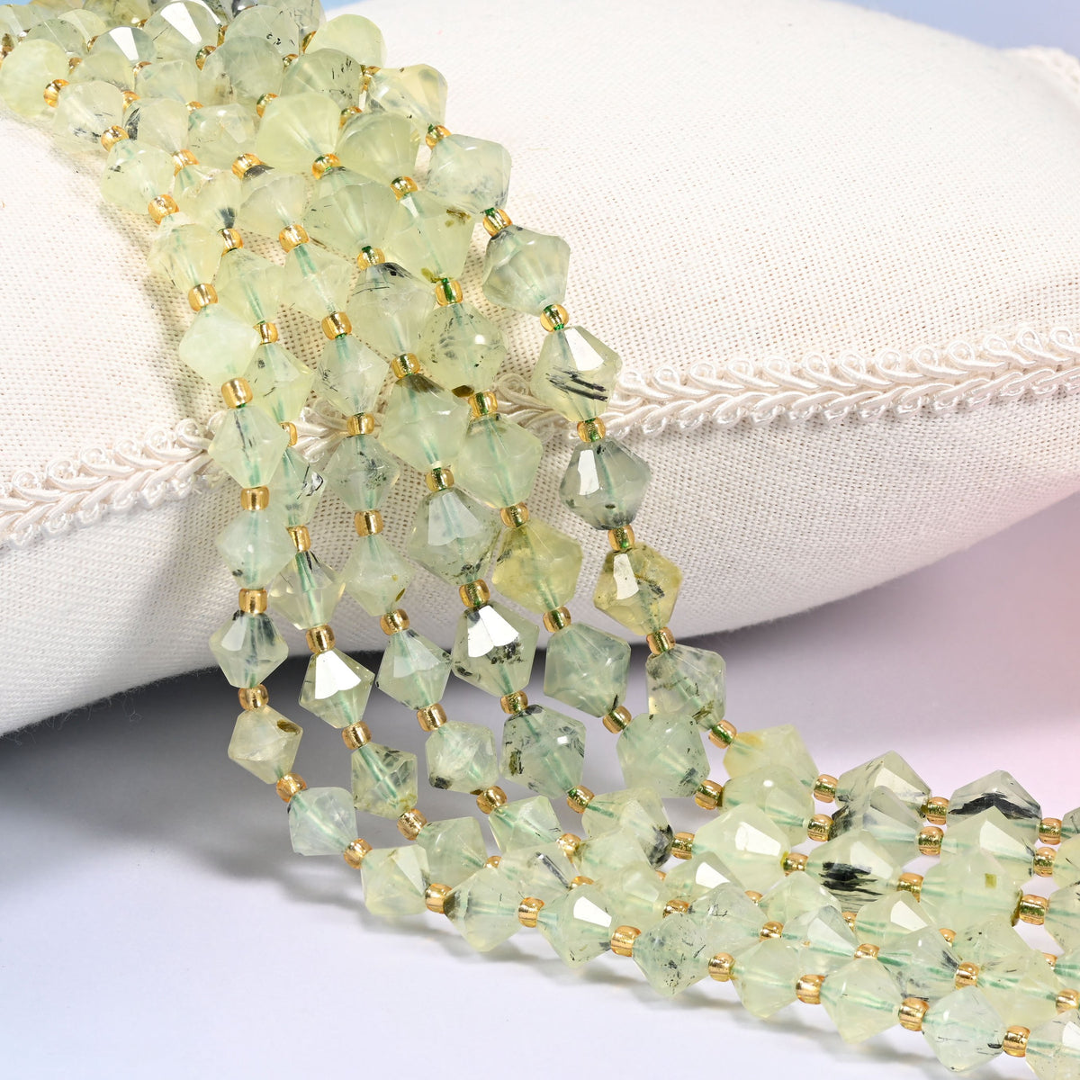Green Prehnite faceted bicone beads, 8mm and 10mm, draped diagonally over a white fabric cushion showing the light green color.