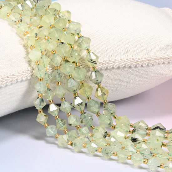 Green Prehnite faceted bicone beads, 8mm and 10mm, draped diagonally over a white fabric cushion showing the light green color.
