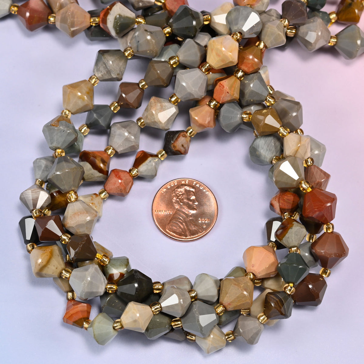 American Picture Jasper faceted bicone beads, 8mm and 10mm, coiled into a circle on a light lavender background, showing the natural stone color variation.