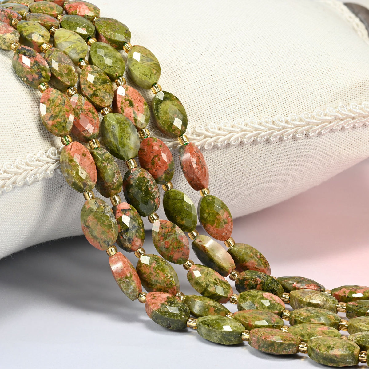 Unakite faceted flat oval beads 9x14mm with small gold seed bead spacers, several strands draped over a white cushion