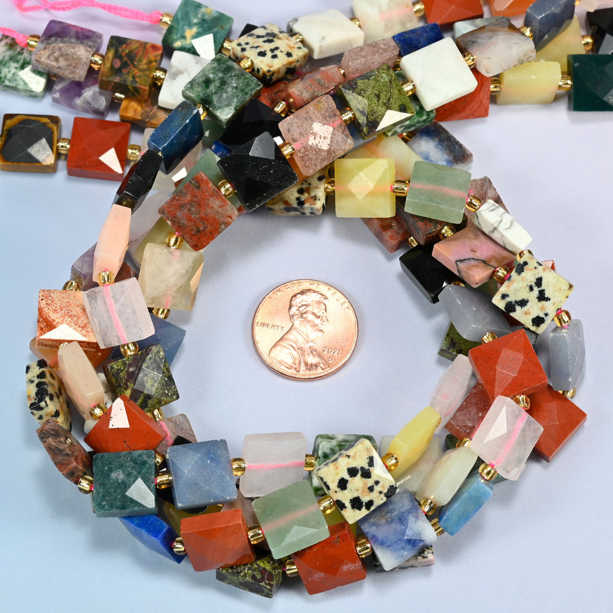 Assorted gemstone faceted flat square beads 10x10mm coiled in a loop around a US penny for scale, close-up of the faceted flat square shape