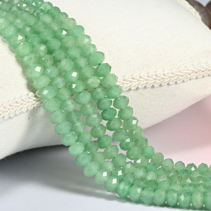 Green aventurine faceted rondelle beads in soft semi-translucent green, three strands draped over a cream linen cushion with braided trim