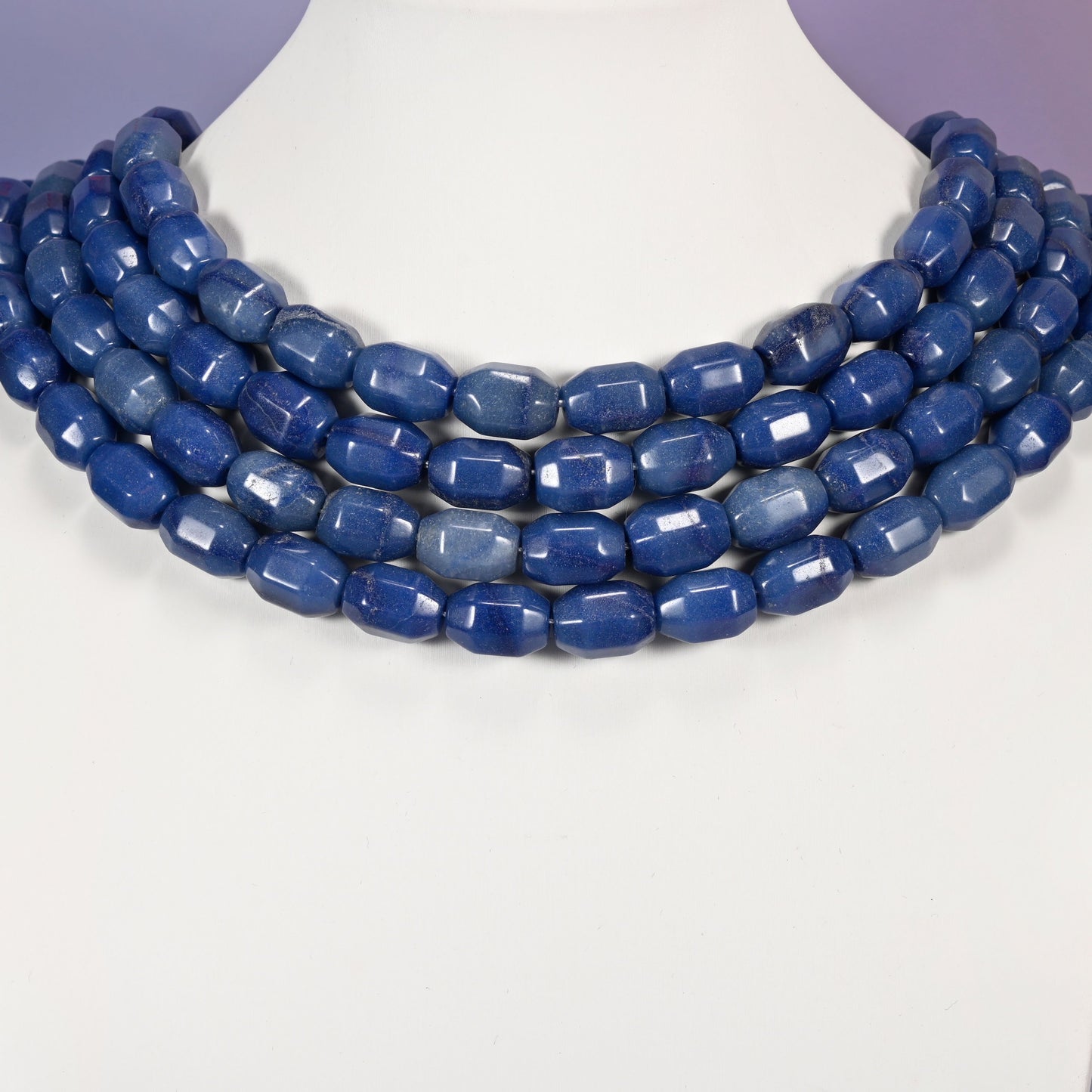 Multi-strand necklace of blue faceted Aventurine barrel beads laid on a white mannequin neck form against a lavender background.
