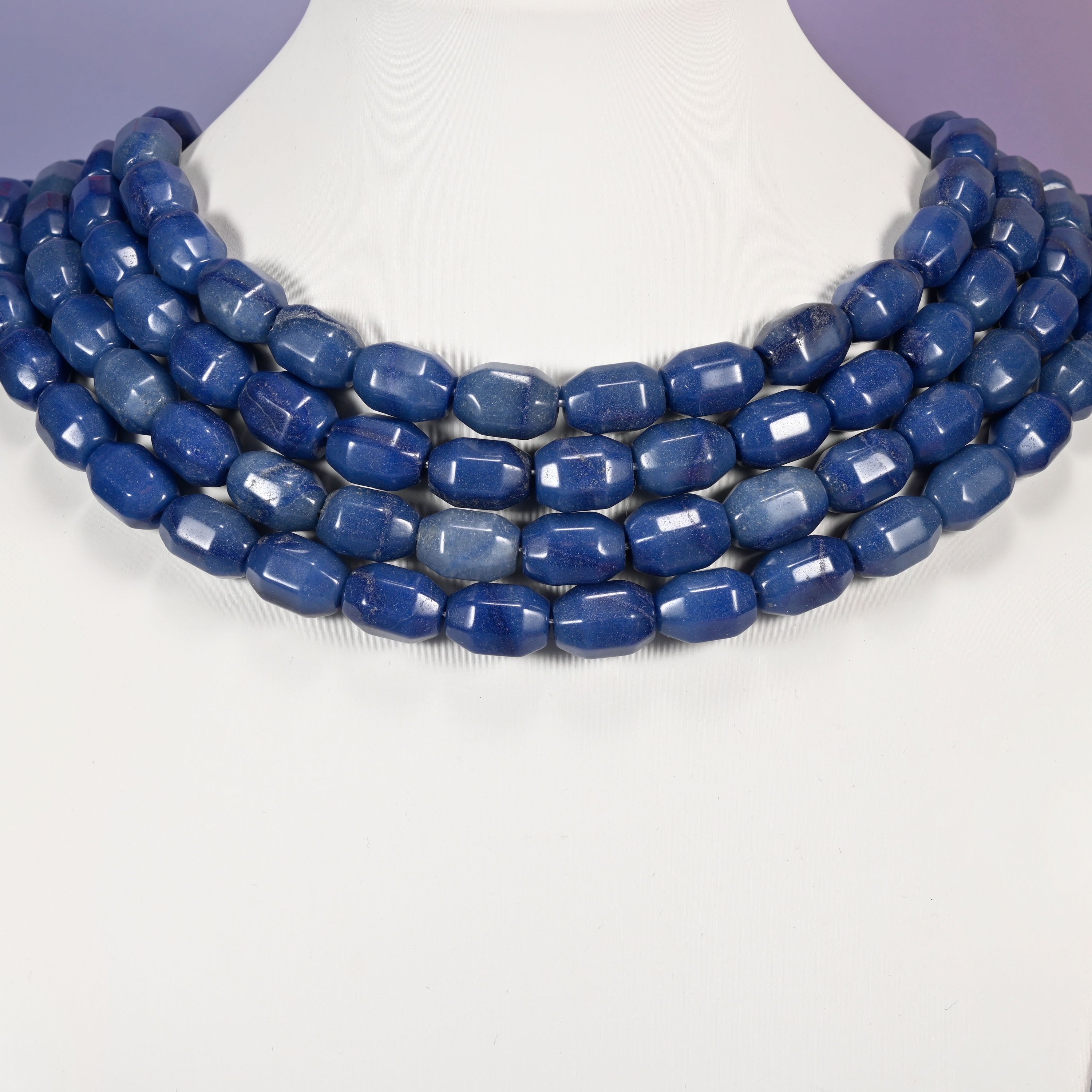 Multi-strand necklace of blue faceted Aventurine barrel beads laid on a white mannequin neck form against a lavender background.
