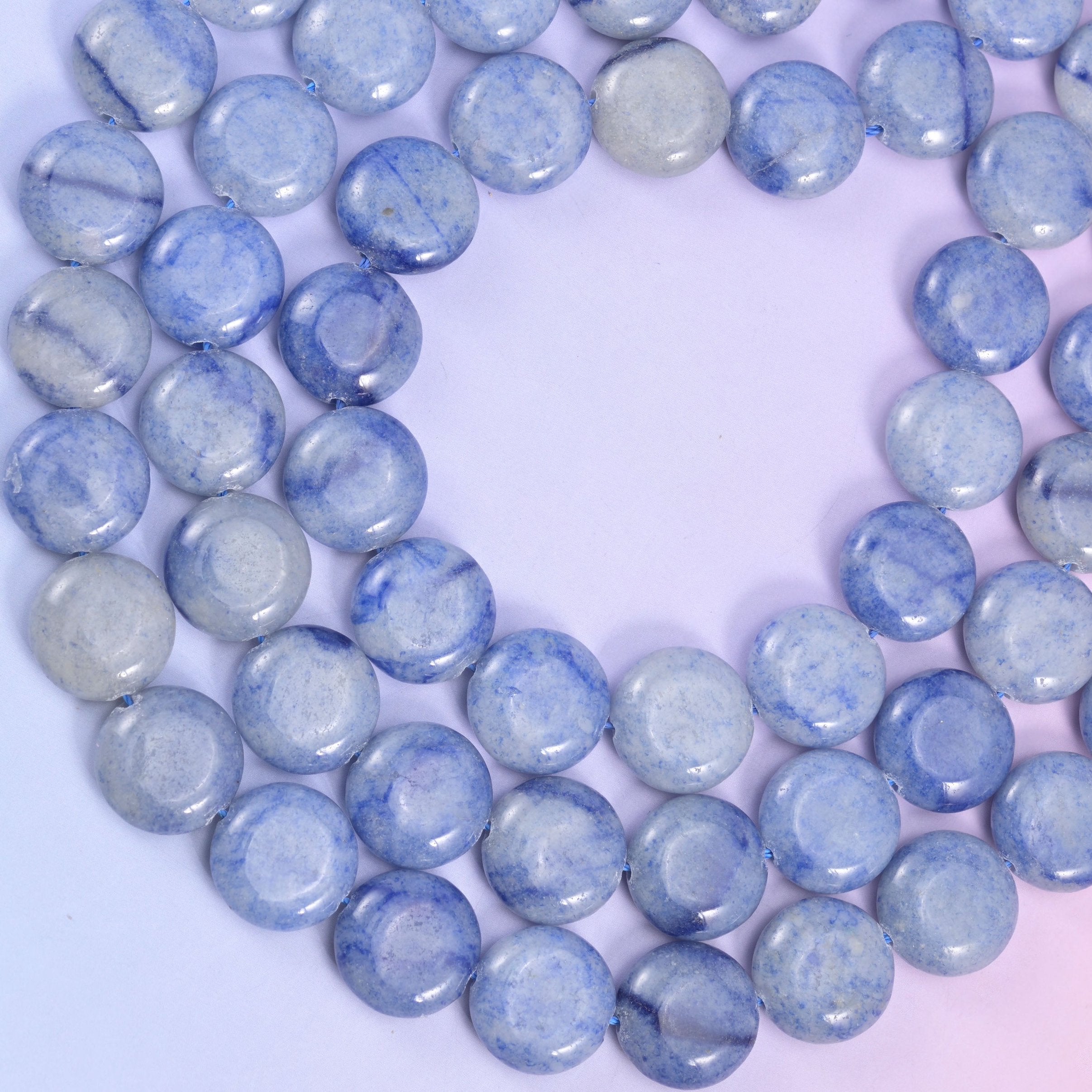 Open loop of blue aventurine coin beads photographed from above on a soft pink-lilac gradient