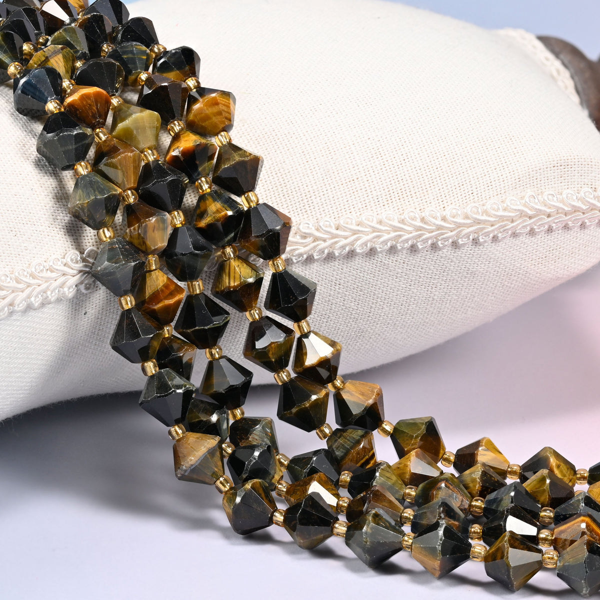 Iron Tiger Eye faceted bicone beads, 10mm, draped diagonally over a white fabric cushion showing the brown and golden banded sheen.