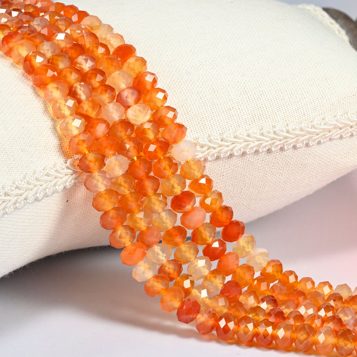 Orange Carnelian faceted rondelle beads draped over a cream fabric cushion, deep red-orange through mid-orange to light peach across the 4x6mm and 5x8mm strands.