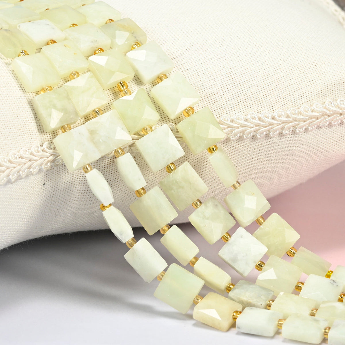 New Jade faceted flat square beads 10x10mm with small gold seed bead spacers, several strands draped over a white cushion