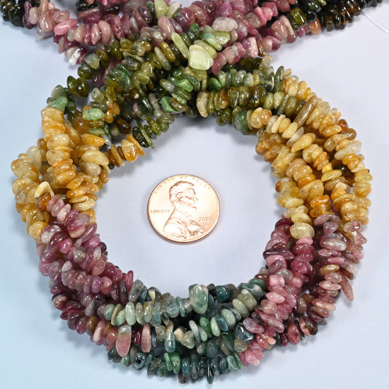 Multicolor tourmaline chip beads 3-8mm coiled in a loop around a US penny for scale, close-up of the small freeform chips
