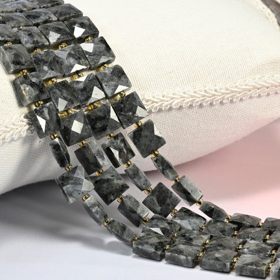 Natural black labradorite faceted flat square beads 10x10mm with small gold seed bead spacers, several strands draped over a white cushion