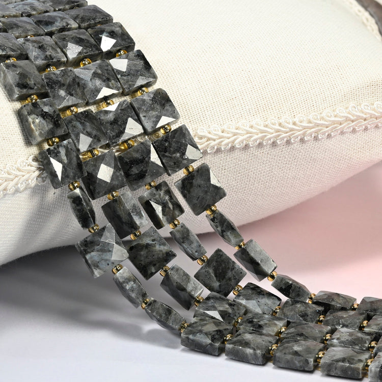 Natural black labradorite faceted flat square beads 10x10mm with small gold seed bead spacers, several strands draped over a white cushion