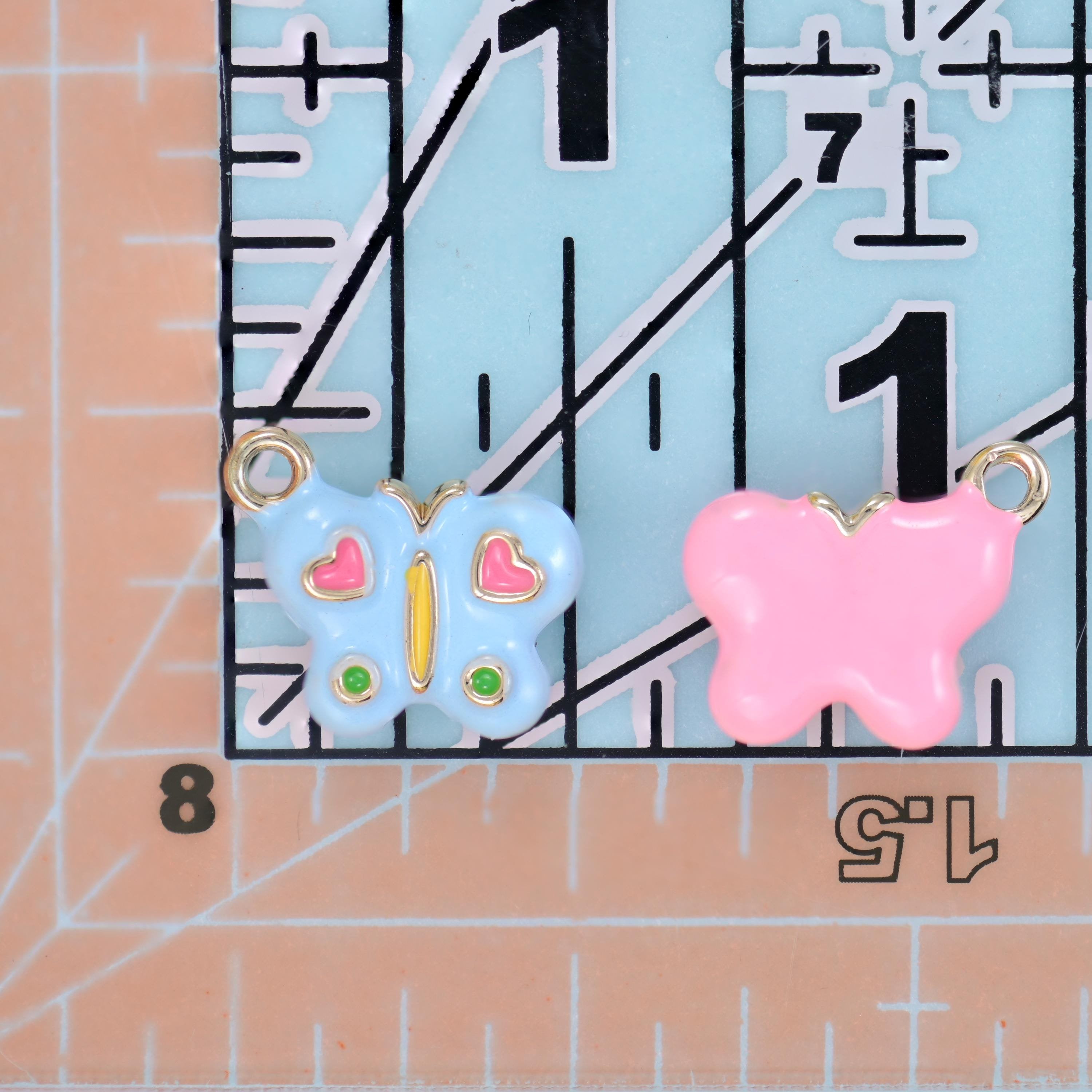 Enamel butterfly charm shown front and back on a ruler, a light blue enamelled face beside a pink piece turned to show its plain reverse.