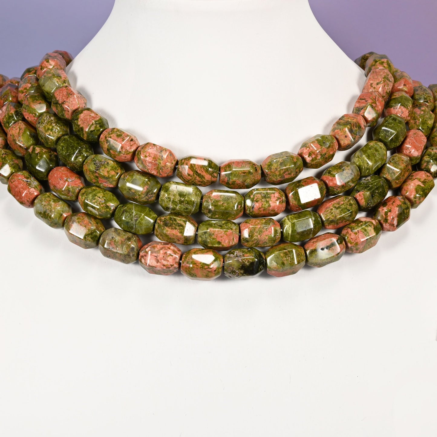 Multi-strand chunky necklace of faceted Unakite barrel beads laid on a white mannequin neck form against a lavender background.