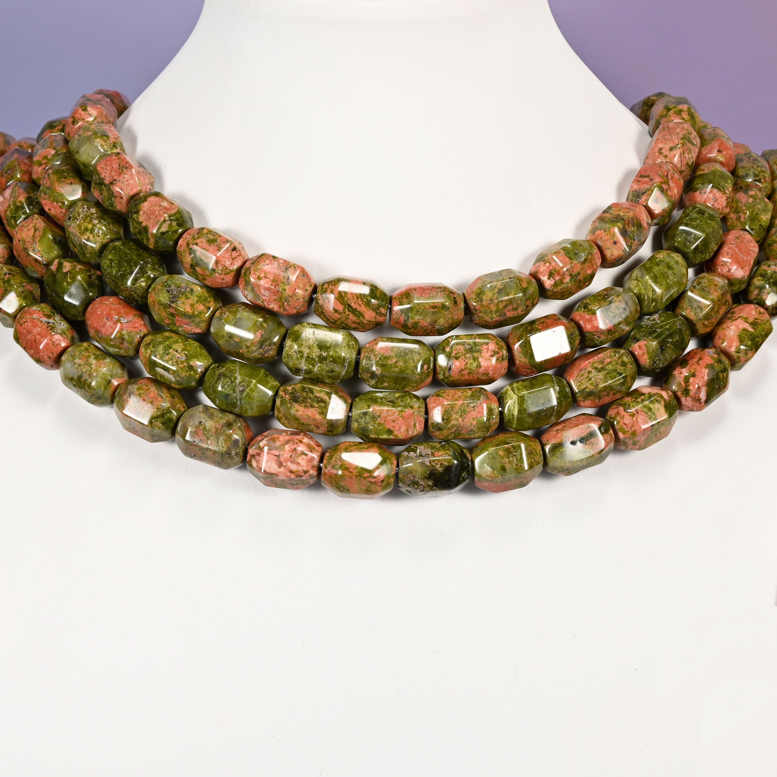 Multi-strand chunky necklace of faceted Unakite barrel beads laid on a white mannequin neck form against a lavender background.