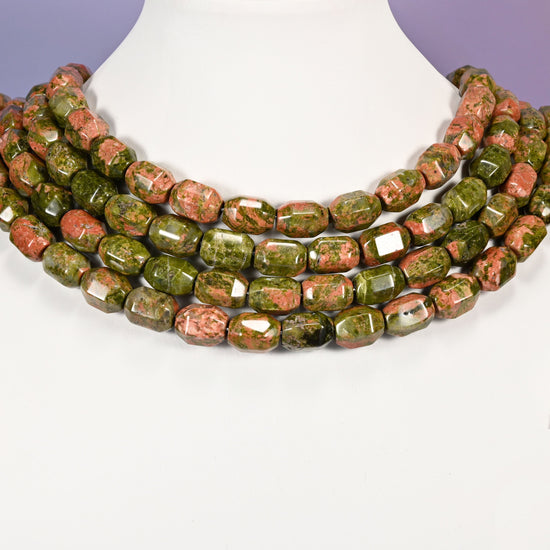 Multi-strand chunky necklace of faceted Unakite barrel beads laid on a white mannequin neck form against a lavender background.