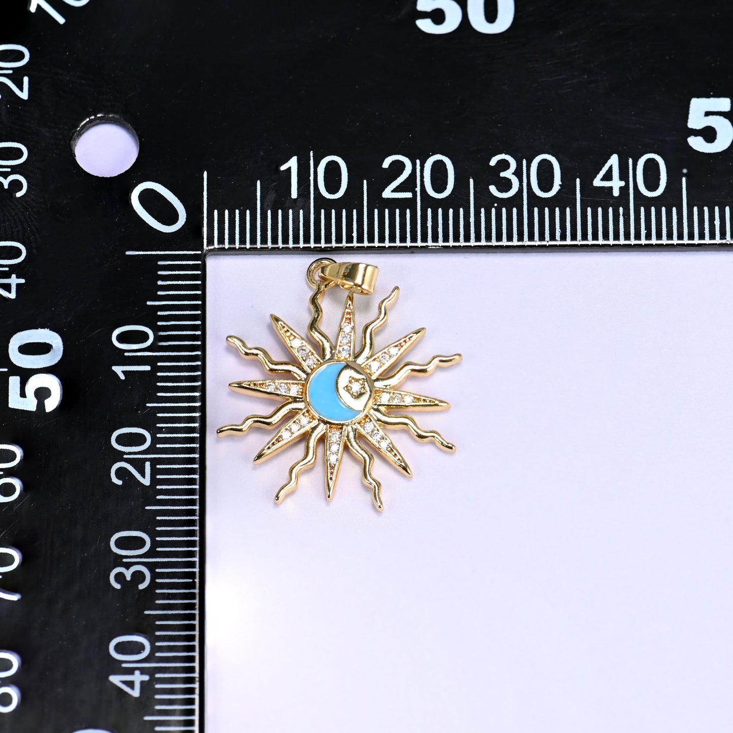 Blue enamel moon sun zirconia charm on a black millimetre ruler, the gold sunburst measuring around 26mm across.