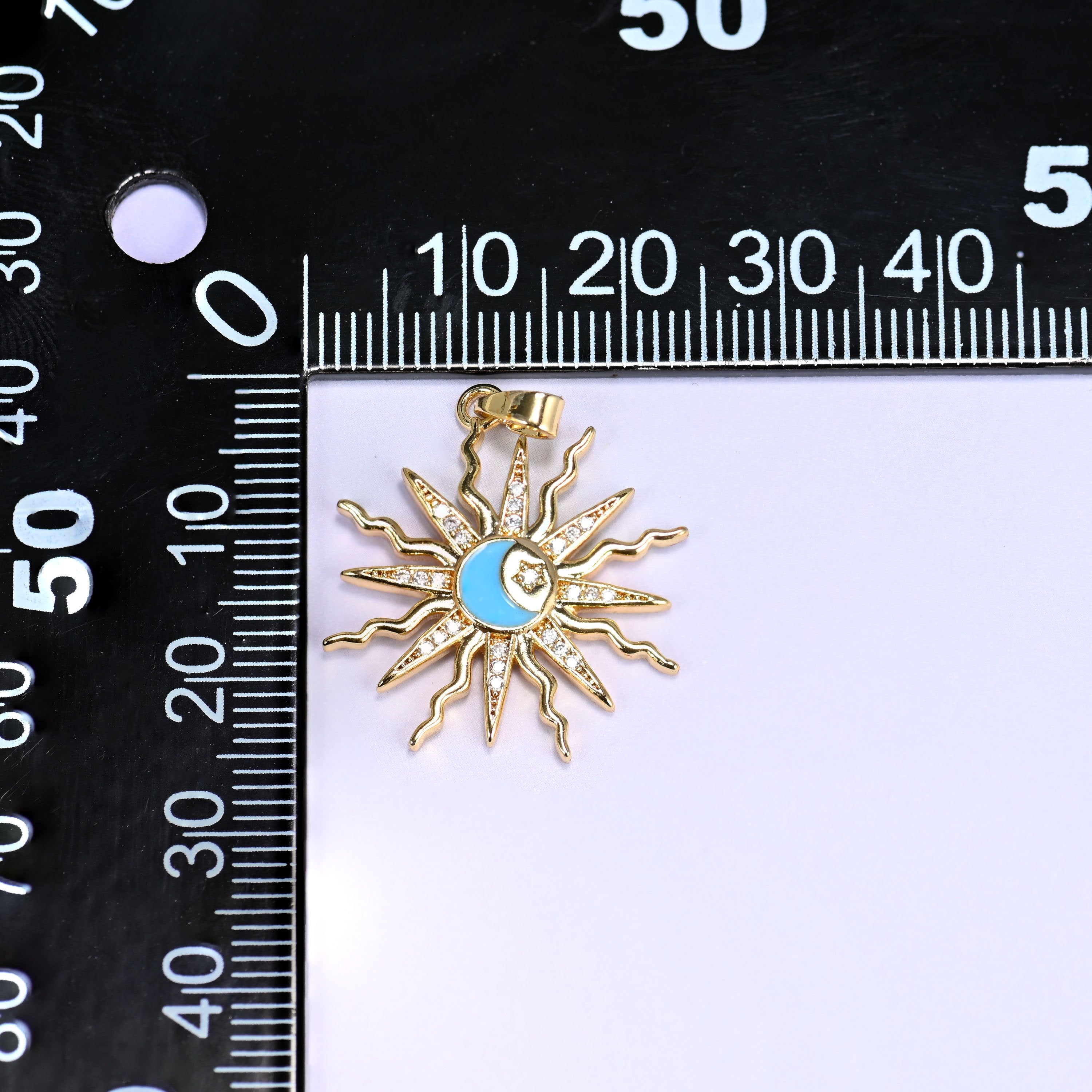 Blue enamel moon sun zirconia charm on a black millimetre ruler, the gold sunburst measuring around 26mm across.