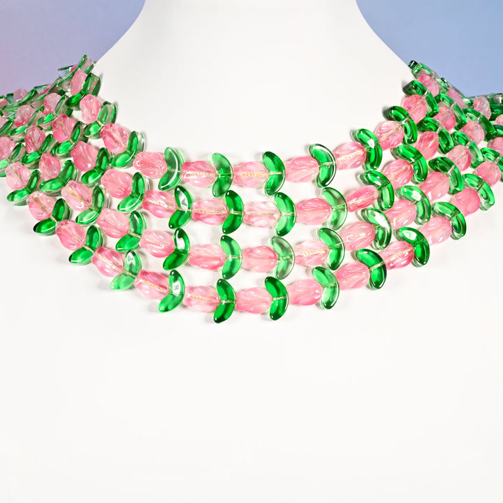 Multi-strand necklace of pink tulip and green leaf glass beads laid on a white mannequin neck form against a lavender background.