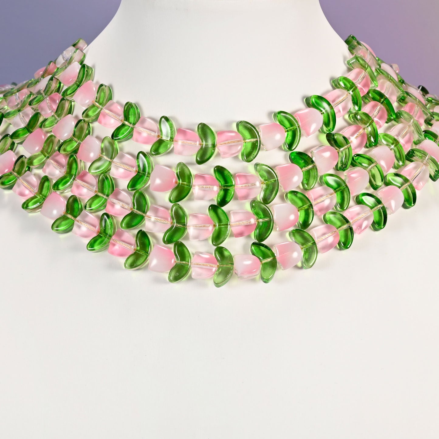 Multi-strand necklace of pink tulip and green leaf glass beads laid on a white mannequin neck form against a lavender background.