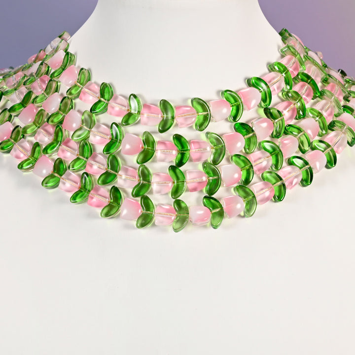 Multi-strand necklace of pink tulip and green leaf glass beads laid on a white mannequin neck form against a lavender background.