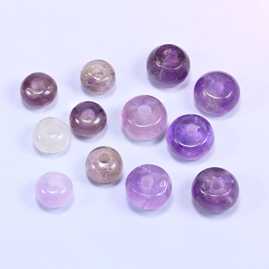 Close-up of individual loose Amethyst beads laid out on a white surface, showing the large center-drilled hole and varied purple coloring of each bead.