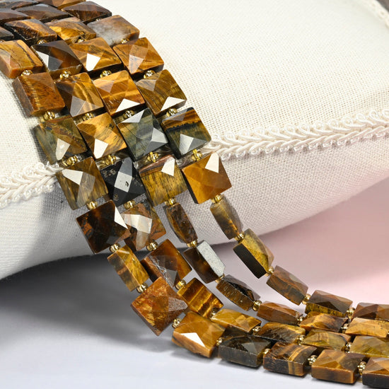 Natural tiger eye faceted flat square beads 10x10mm with small gold seed bead spacers, several strands draped over a white cushion