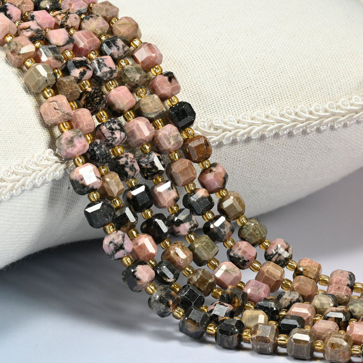 Natural rhodonite faceted cube beads in pink, tan and black, strands draped over a white linen cushion with gold glass seed bead spacers