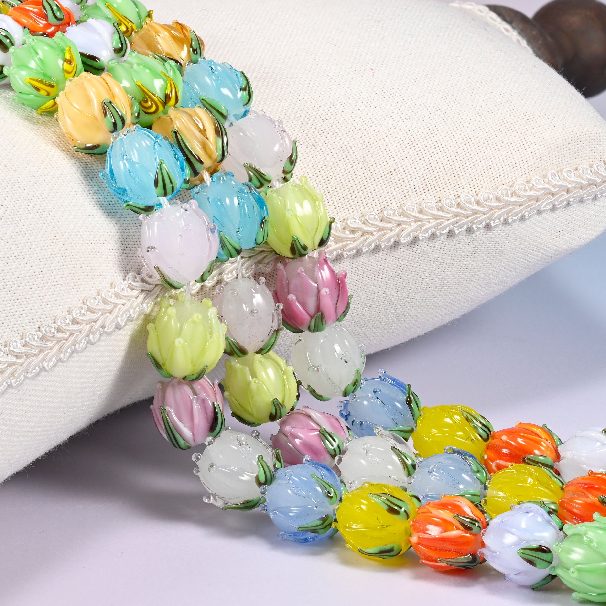 Close-up of multicolor tulip bud lampwork glass beads cascading diagonally over a cream woven pillow with decorative trim, a dark wooden object visible in the corner.