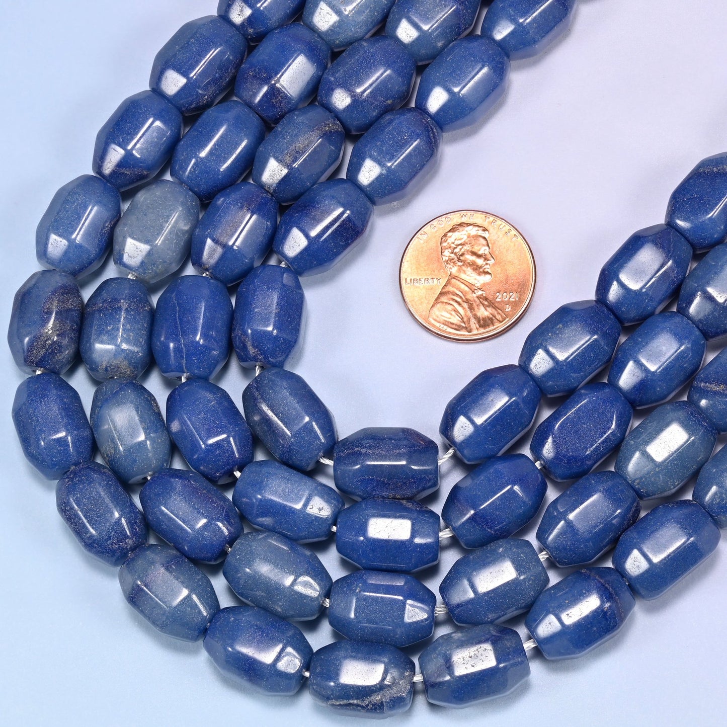 Circular coil of blue faceted Aventurine barrel bead strands on a light blue background with a penny in the center for size comparison.