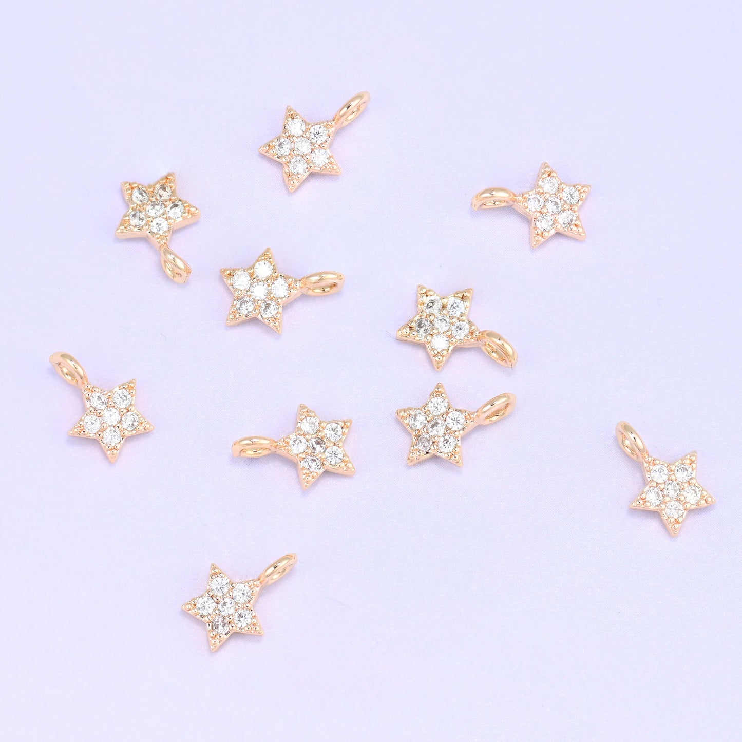 Small gold charms, nine 18K gold plated star charms with clear cubic zirconia scattered on a pale backdrop