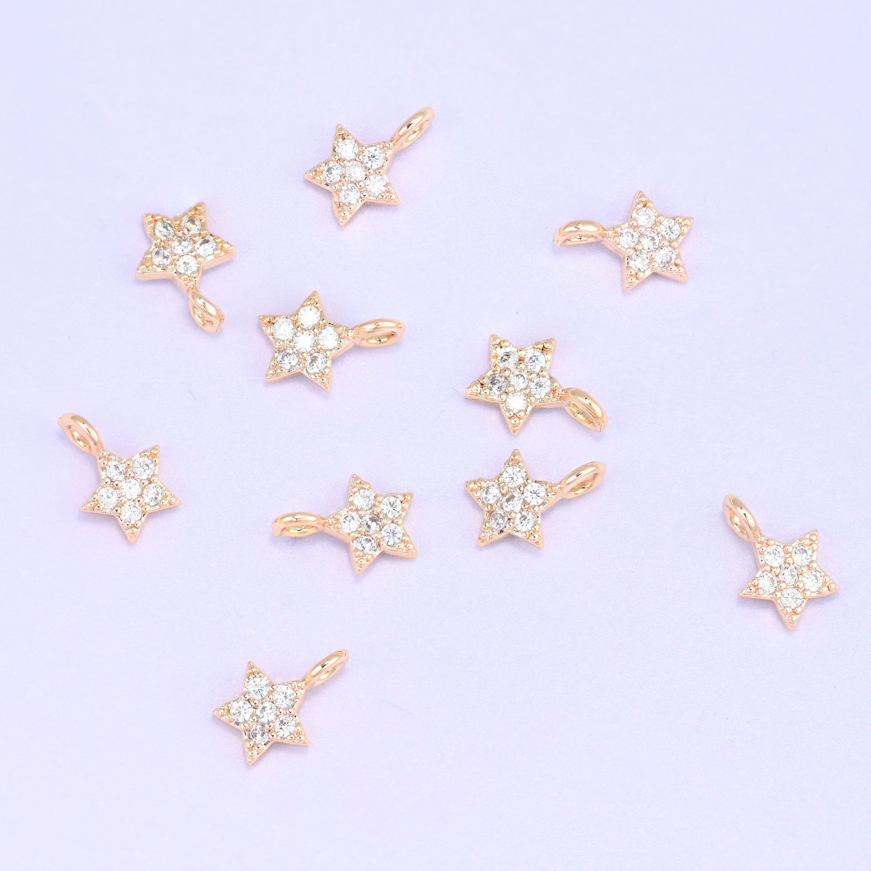 Small gold charms, nine 18K gold plated star charms with clear cubic zirconia scattered on a pale backdrop
