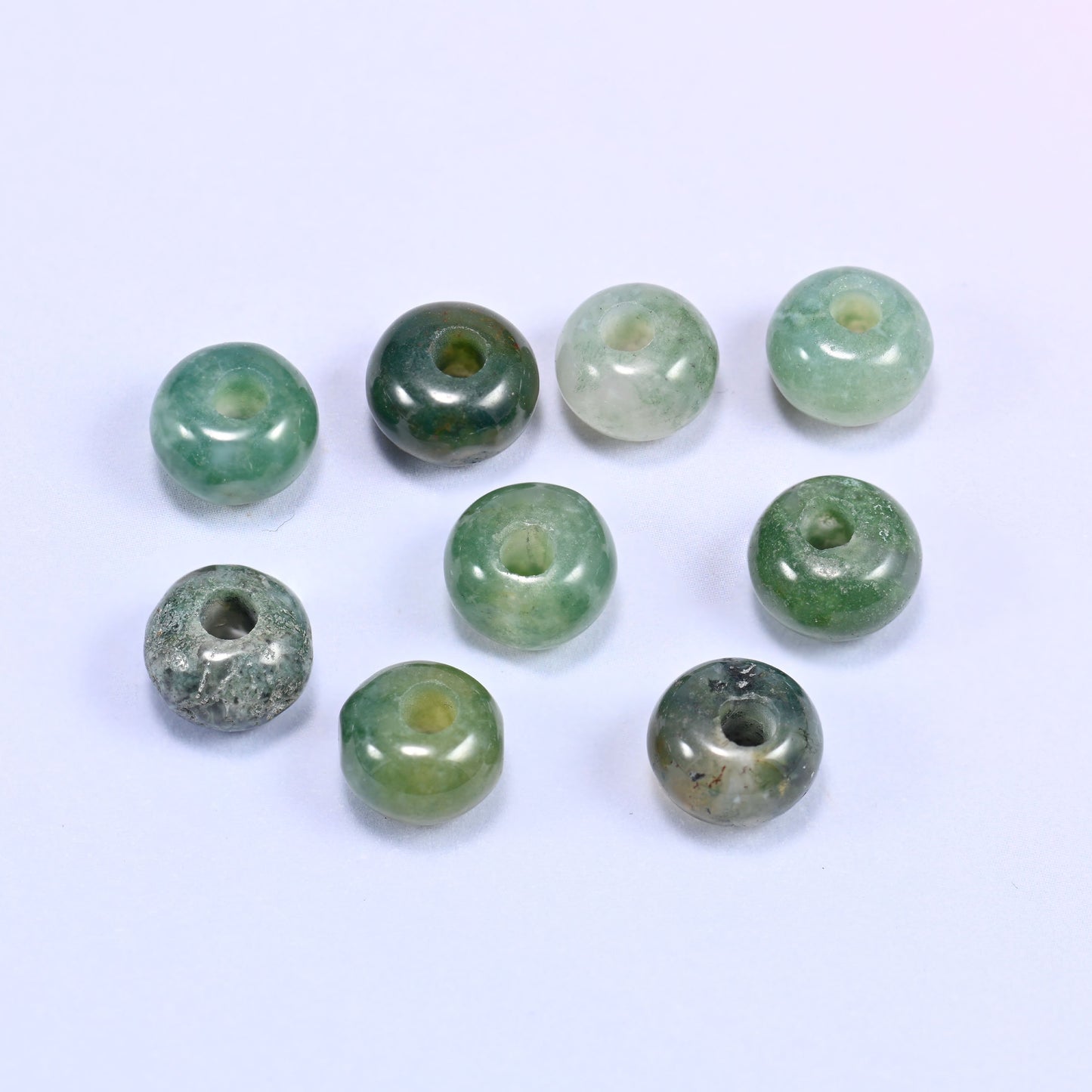 Close-up of individual loose Moss Agate beads laid out on a white surface, showing the large center-drilled hole and translucent green coloring of each bead.