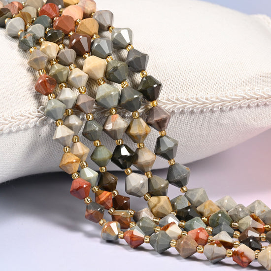 American Picture Jasper faceted bicone beads, 8mm and 10mm, draped diagonally over a white fabric cushion showing the multicolor scenic banding.