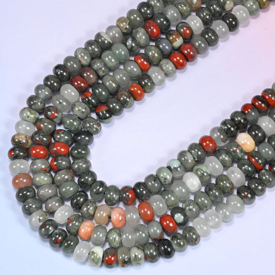 Overhead view of several rows of African Bloodstone round beads showing the mix of gray-green, red, and cream coloring on a light lavender surface.